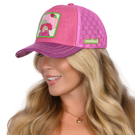 Woman wearing magenta linen trucker cap hat with Strawberry Shortcake Raspberry Tart woven patch against a white background
