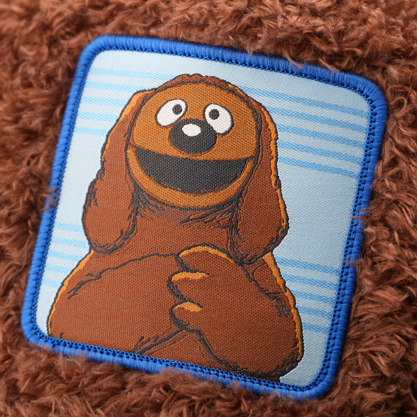 Close-up of a woven patch showing Rowlf the Dog from The Muppets smiling with hands clasped on a light blue striped background, stitched onto fuzzy-textured brown fabric with a blue border.