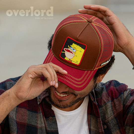 Man wearing brown and maroon OVERLORD X SpongeBob Caveman SpongeBob meme trucker baseball cap hat with yellow zig zag stitching. PVC Overlord logo.