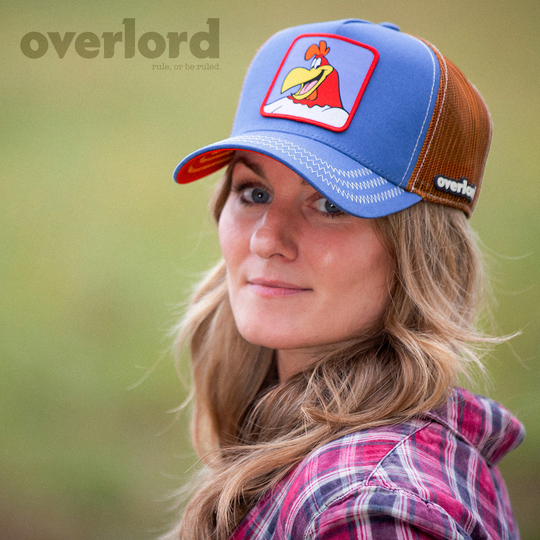 Woman wearing blue OVERLORD X Looney Tunes smiling Foghorn trucker baseball cap with cream stitching. PVC Overlord logo.