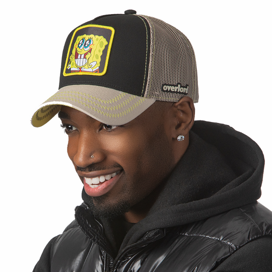 Man wearing black and tan OVERLORD X SpongeBob Big Smile SpongeBob trucker baseball cap hat with yellow zig zag stitching. PVC Overlord logo.