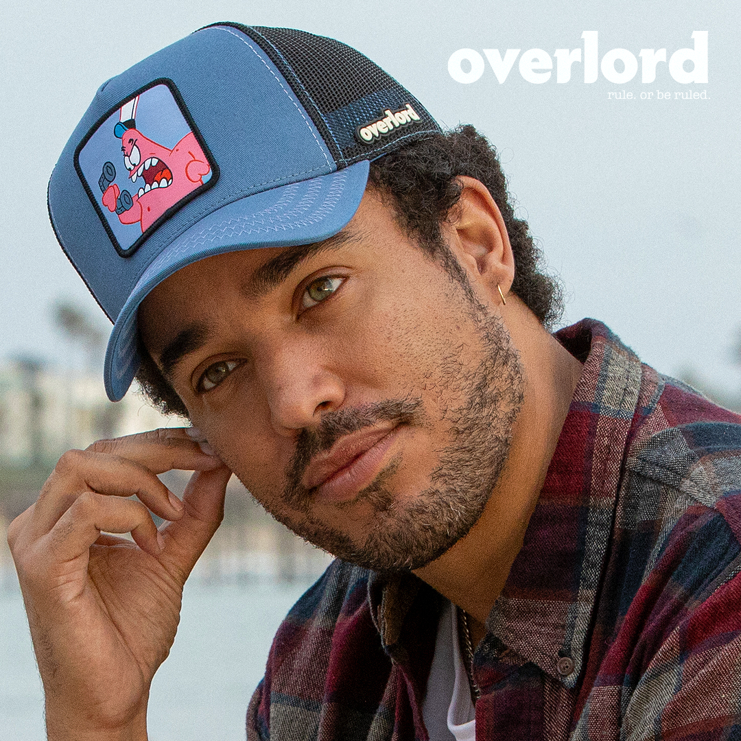 Man wearing blue OVERLORD X SpongeBob Patrick yelling at phone trucker baseball cap hat with light blue zig zag stitching. PVC Overlord logo.