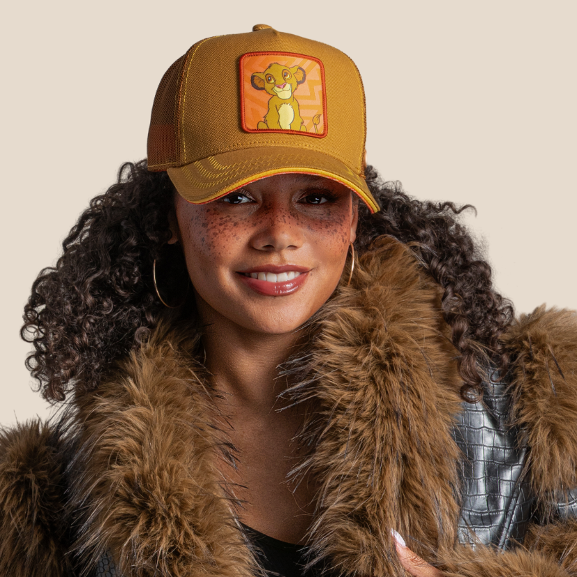 Model wearing the tan young Simba from Disney's The Lion King retro trucker cap with a red-trimmed brim and brown mesh side.