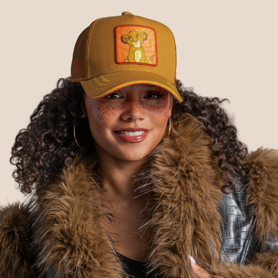 Model wearing the tan young Simba from Disney's The Lion King retro trucker cap with a red-trimmed brim and brown mesh side.