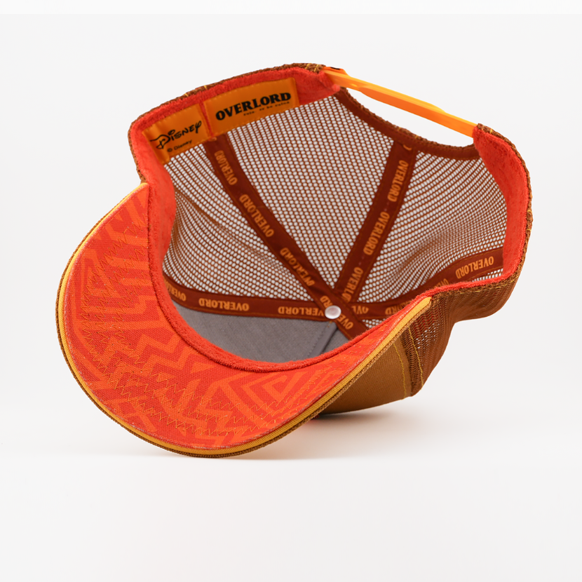 Underside of the young Simba from Disney's The Lion King retro trucker cap showing an orange patterned brim, brown mesh crown, Disney tag, and orange Overlord branded taping with brown text.