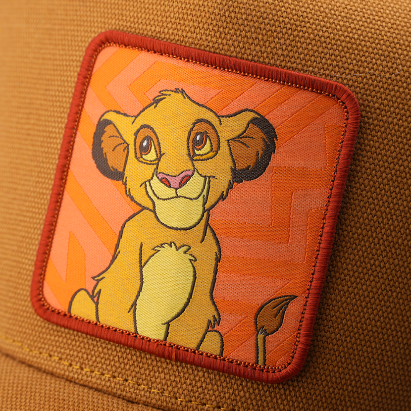 Close-up woven patch of young Simba from Disney's The Lion King smiling, on an orange patterned background with a red border, stitched onto a tan corduroy-textured retro trucker cap.