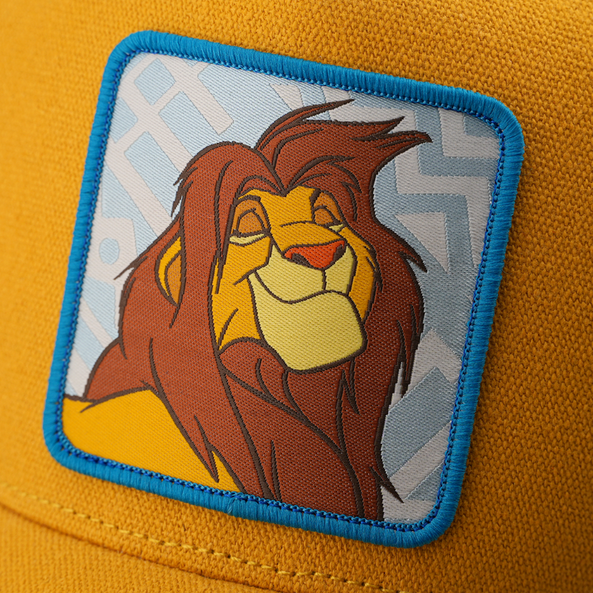 Close-up woven patch of adult Simba from Disney's The Lion King with eyes closed in contentment, on a light blue patterned background with a blue border, stitched onto a yellow corduroy-textured retro trucker cap.
