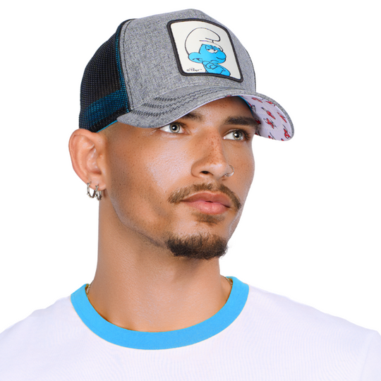 Man wearing heather gray trucker cap with black mesh featuring a woven patch of Grouchy Smurf from the Smurfs attached to crown