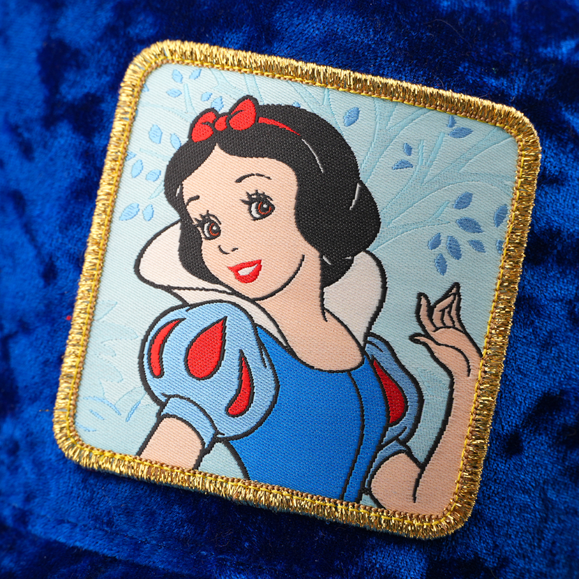 Woven patch of Snow White from Disney's Snow White and the Seven Dwarfs smiling against a leafy background, with a gold border, stitched onto the blue velvet retro trucker cap.