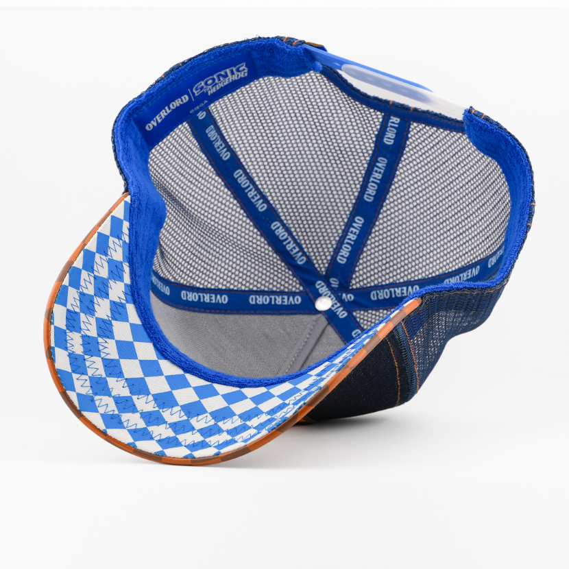 Underside of the Sonic the Hedgehog retro trucker cap, showing a blue and white argyle-pattern brim, Sonic the Hedgehog tag, grey mesh crown, and blue Overlord branded taping with white text.
