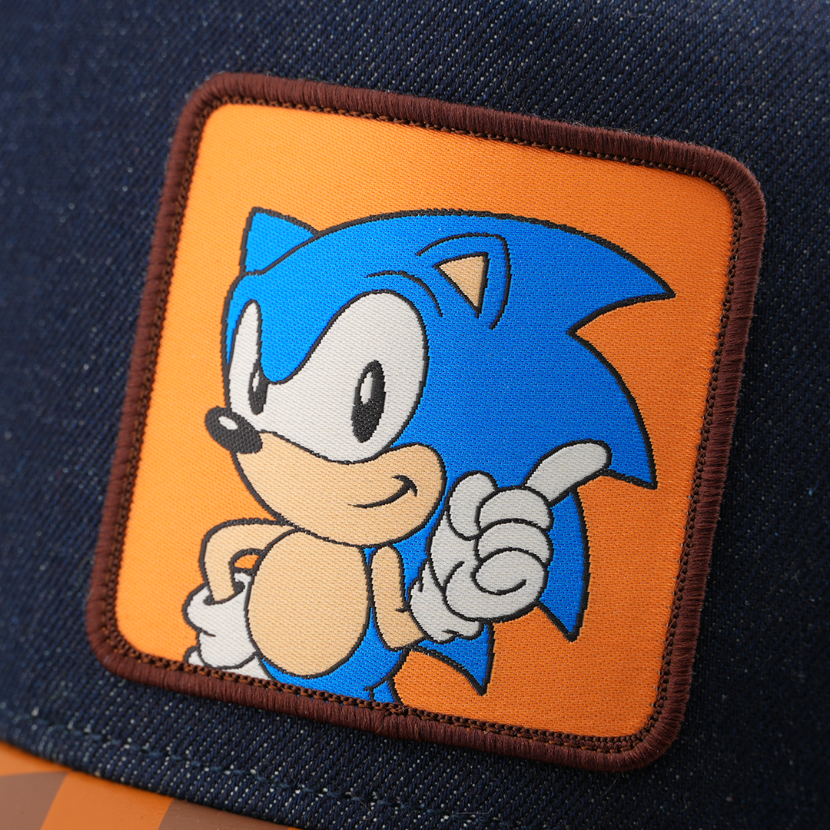 Woven patch of Sonic the Hedgehog pointing forward, set against an orange background with dark brown border, stitched onto the denim-textured navy retro trucker cap.