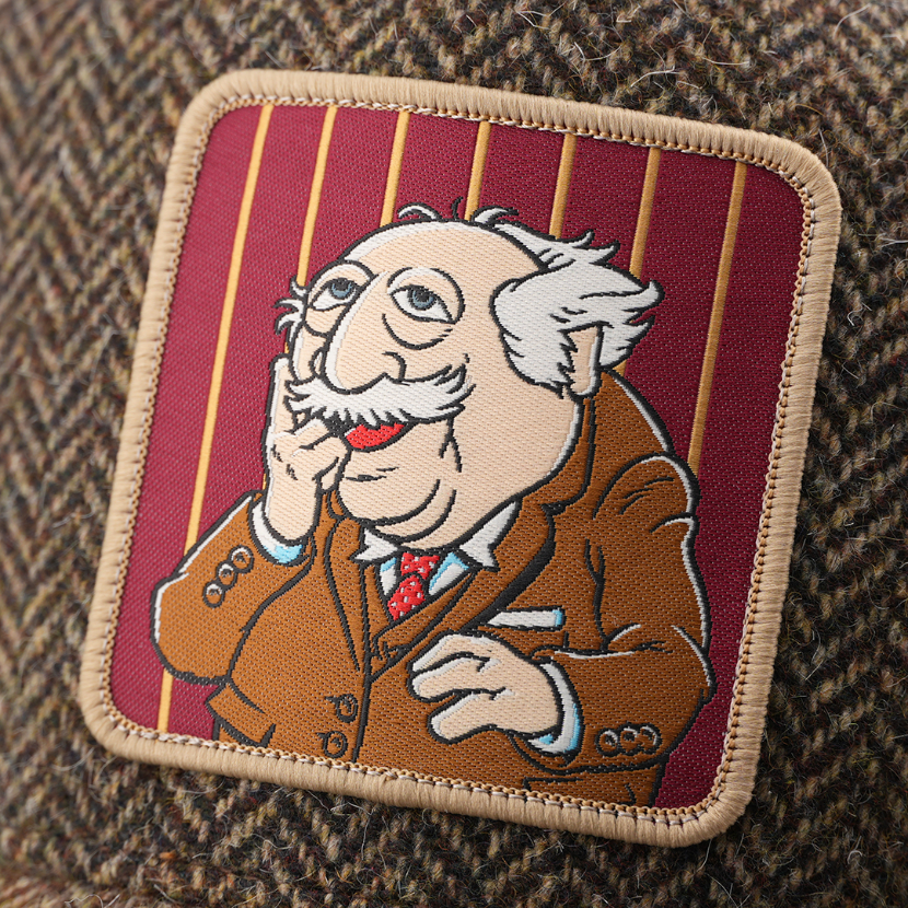 Close-up of a woven patch showing Statler from The Muppets in a brown suit with a red tie on a maroon striped background, stitched onto tweed-textured brown fabric with a tan border.