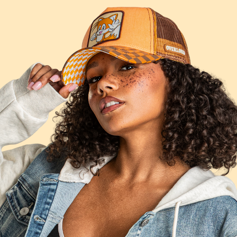 Model wearing the orange Tails from Sonic the Hedgehog retro trucker cap, with checkered brim and brown mesh sides.