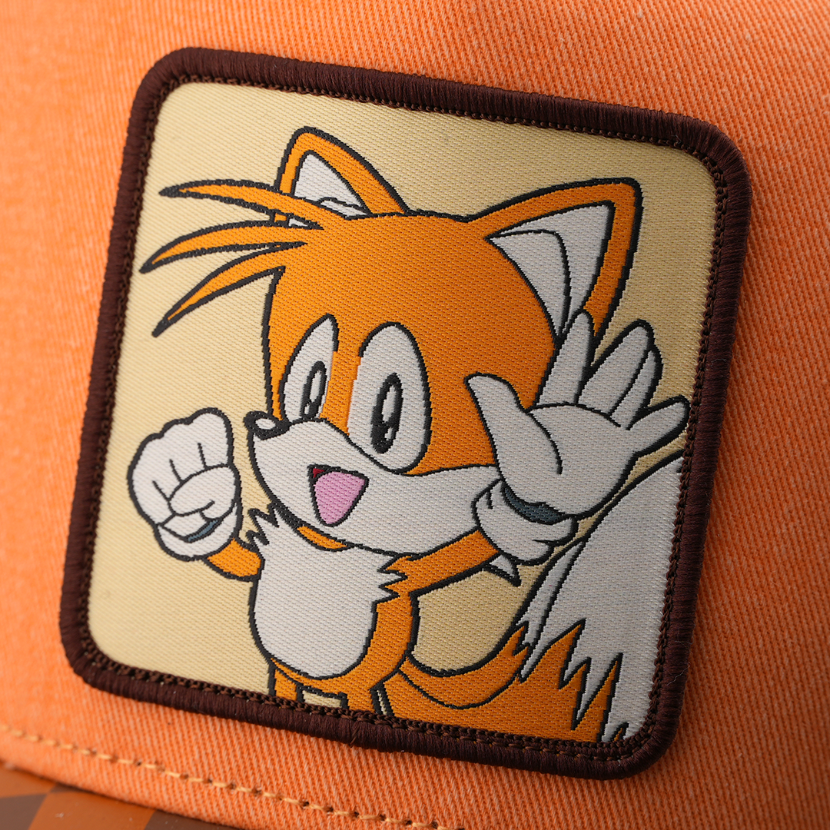 Woven patch of Tails from Sonic the Hedgehog in a dynamic pose with raised hands, set against a cream background with dark brown border, stitched onto the orange retro trucker cap.