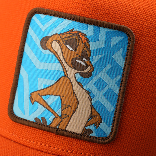 Close-up woven patch of Timon from Disney's The Lion King in a confident pose, on a blue patterned background with a brown border, stitched onto an orange corduroy-textured retro trucker cap.