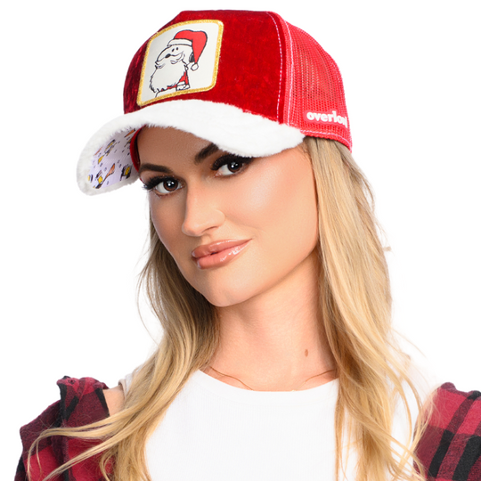 Woman wearing red crushed velvet trucker cap with white faux fur brim and red mesh featuring a woven patch of Snoopy from the Peanuts wearing a Santa suit attached to the crown.